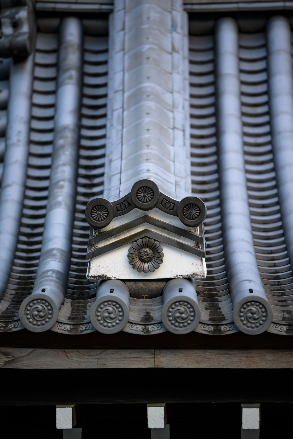 A traditional tiled Japanese roof is shown from the side, with the tile layers swooping down in a steep curve, ending with cast reliefs of traditional heraldic symbols, kamon, associated with the imperial family. Four tile end caps, or gatō, have the three-part 'tomoe' symbol surrounded by a circle of dots. A single cast relief at the end of a primary tile cap featured in the center of the photo is a chrysanthemum in full bloom with three stylized chrysanthemum 'kiku' kamon, the Imperial Seal of Japan, in a wide triangular pattern above it. The roof tiles have a pronounced slate blue color and the wooden support beams are barely visible in the shadows beneath the tiles.