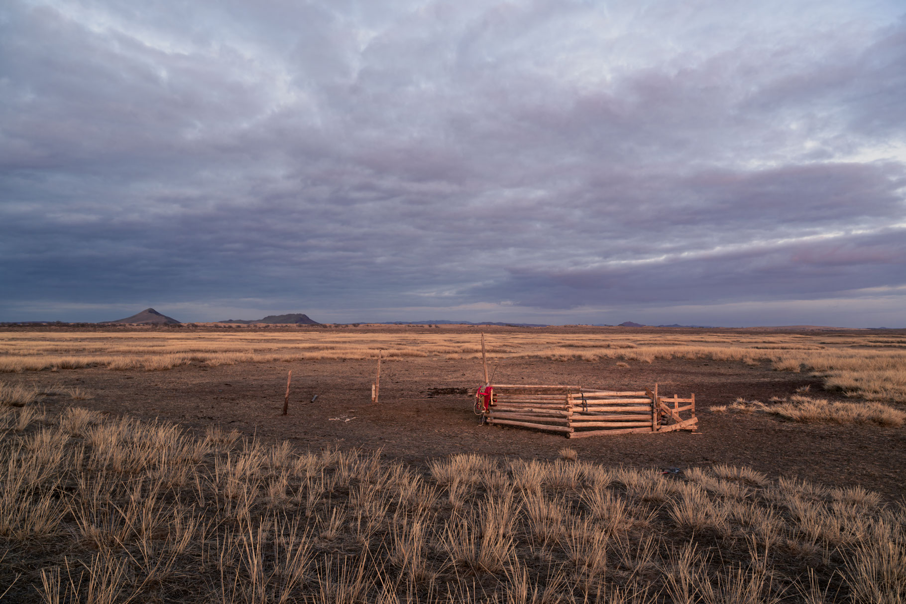 A broad open plain with stiff, tall brown grasses extends out to the horizon which is broken only by a few small, distant hills. The sky is filled with a thick layer of darkening clouds and the light is turning golden as the sun sets somewhere behind the vantage point of the camera. In the center of the field directly ahead of the camera is a small clearing with a rough wooden paddock, empty of any livestock. A single, red saddle from a camel is draped over the paddock railings.