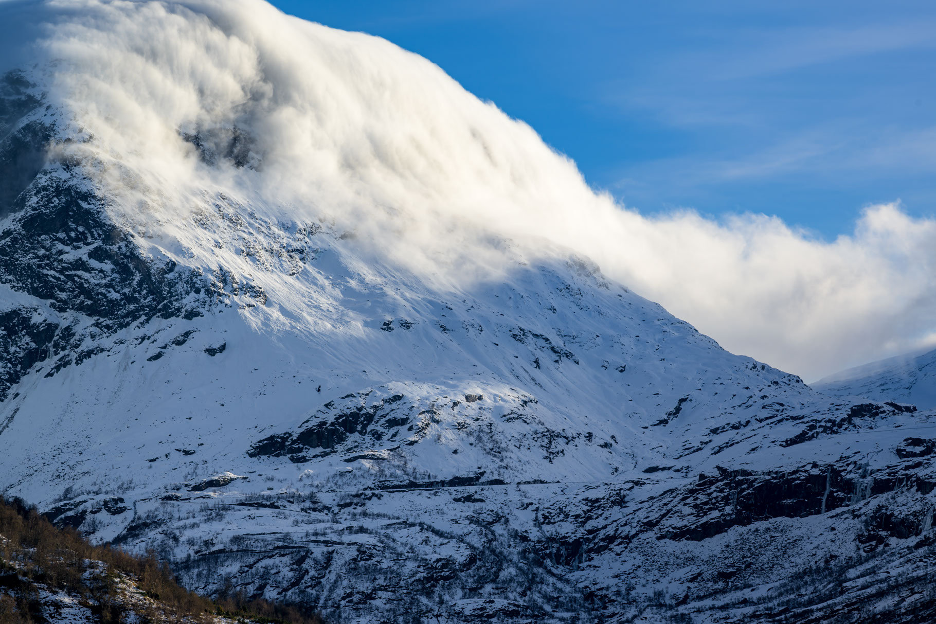 A snow covered mountain dominates the frame, with a blue sky visible just in the upper right. A smaller mountain is barely visible in the bottom left sparsely populated by pine trees. The large mountain peak featured in the image extends upward and to the left across the entire photo, but is covered by clouds in its top third. The clouds are being pushed over the top of the mountain by the wind, revealing the snow covered rocks beneath. Much of the mountain is in shadow, but the sun is directly illuminating the still-cloudcovered portion.