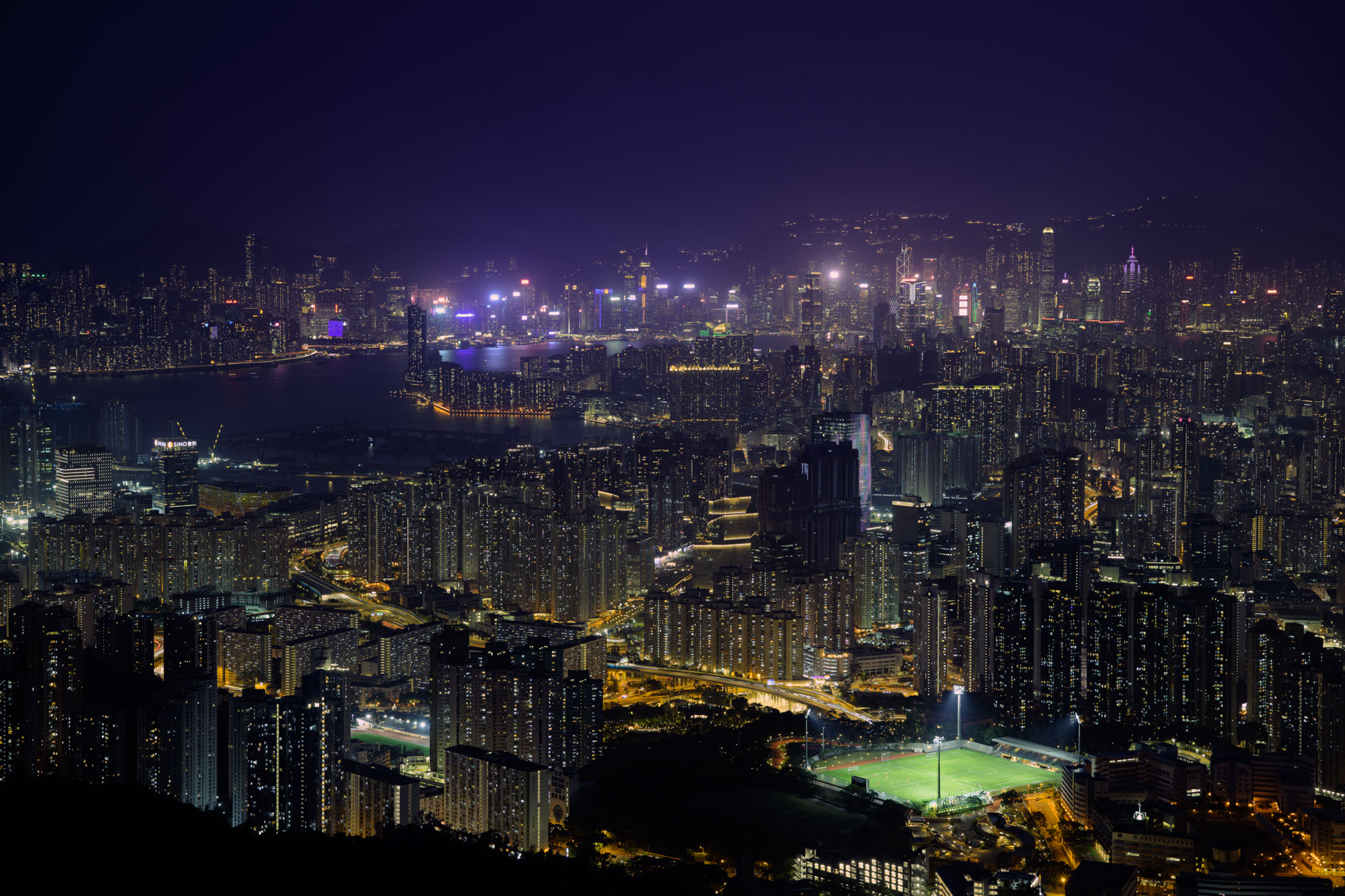 A dense city at night is full of apartment and office towers, with warm yellow street lights and thousands of interior lights shining through windows filling the bottom two thirds of the image. A bay is visible in the background, lined with tall buildings, giant bright billboards and signs visible on the tops of the buildings on the far side of the water. In the distance, just barely visible, are the outlines of hills surrounding the city, dotted with the lights of homes. No stars are visible in the deep purple haze of the clouded sky which reflects back the light of the city. In the bottom right of the photo is a brightly illuminated sports field, home to two soccer pitches with the tiny figures of yellow-uniformed players stopped in streaked motion from the slow shutter speed of the camera.