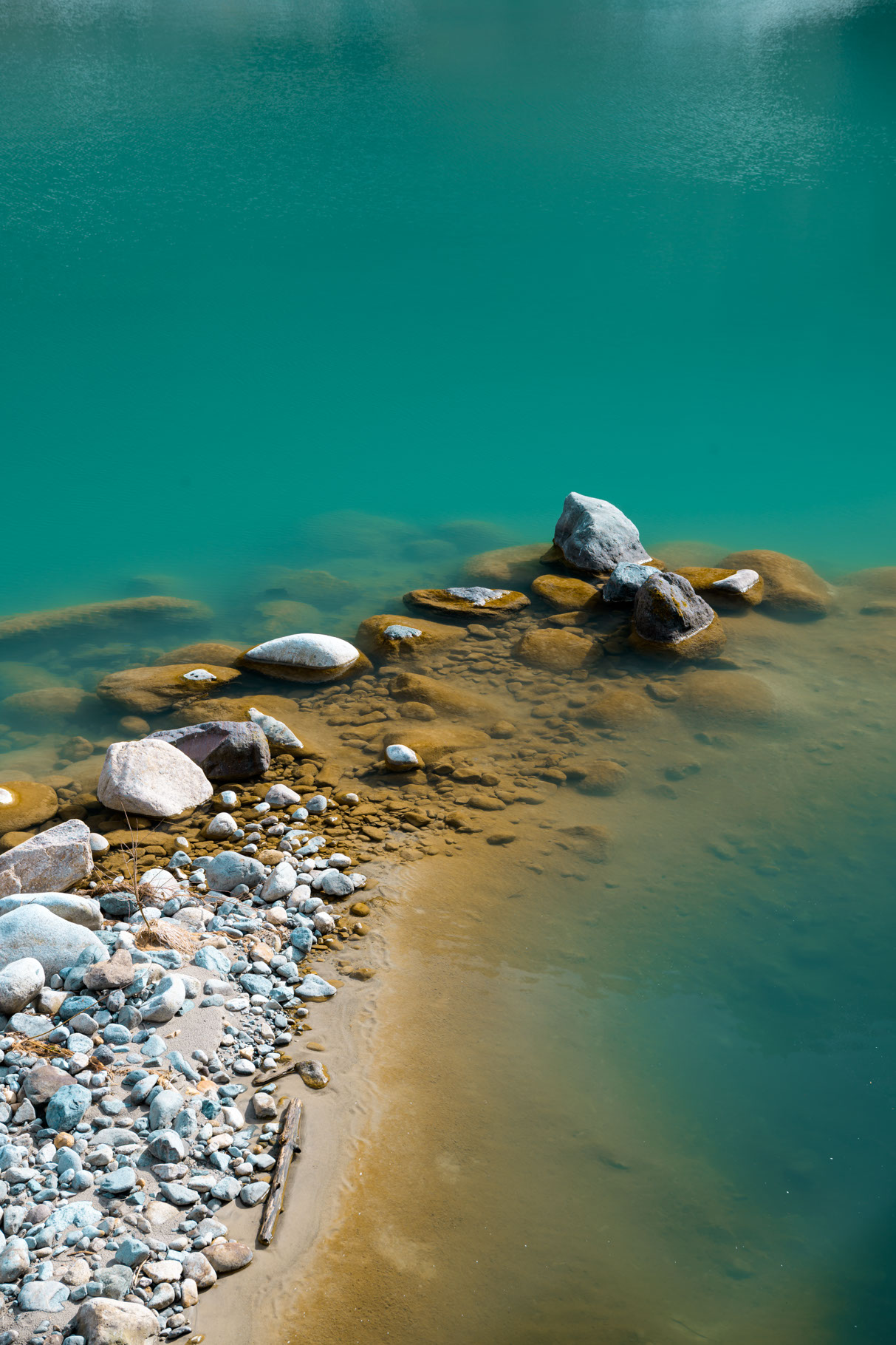 A thin jut of light colored sand extends upward and to the right from the lower left corner of the frame into a clear body of water. The water is a deep blue-green and the sand beneath the surface appears tinted a rich ochre because of the hue of the water. Covering much of the sand, both above and below the water, are large and small rocks, most smoothed by the water over time, but some of the larger ones retaining a rougher surface and sharper edges. A small, straight piece of bleached driftwood sits just above the water line on the sand near the bottom of the photo. The end of the sand bar is dominated by larger rocks, almost all fully submerged, but a few of the largest stick up out of the water, their exposed surfaces completely dried by the day's sun.