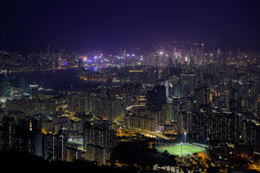 A dense city at night is full of apartment and office towers, with warm yellow street lights and thousands of interior lights shining through windows filling the bottom two thirds of the image. A bay is visible in the background, lined with tall buildings, giant bright billboards and signs visible on the tops of the buildings on the far side of the water. In the distance, just barely visible, are the outlines of hills surrounding the city, dotted with the lights of homes. No stars are visible in the deep purple haze of the clouded sky which reflects back the light of the city. In the bottom right of the photo is a brightly illuminated sports field, home to two soccer pitches with the tiny figures of yellow-uniformed players stopped in streaked motion from the slow shutter speed of the camera.